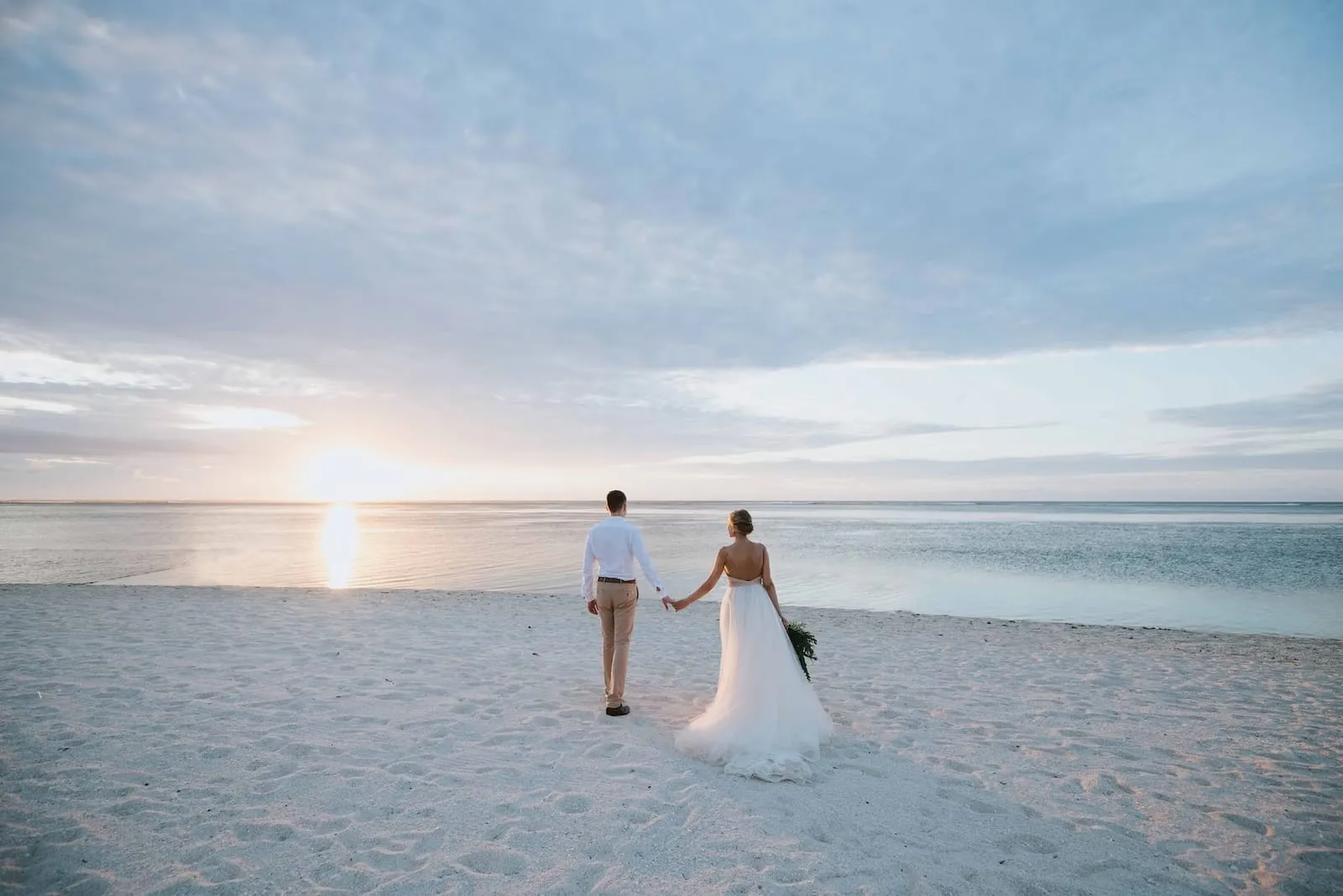 the back of a couple walking on an empty white sand beach with the sun setting